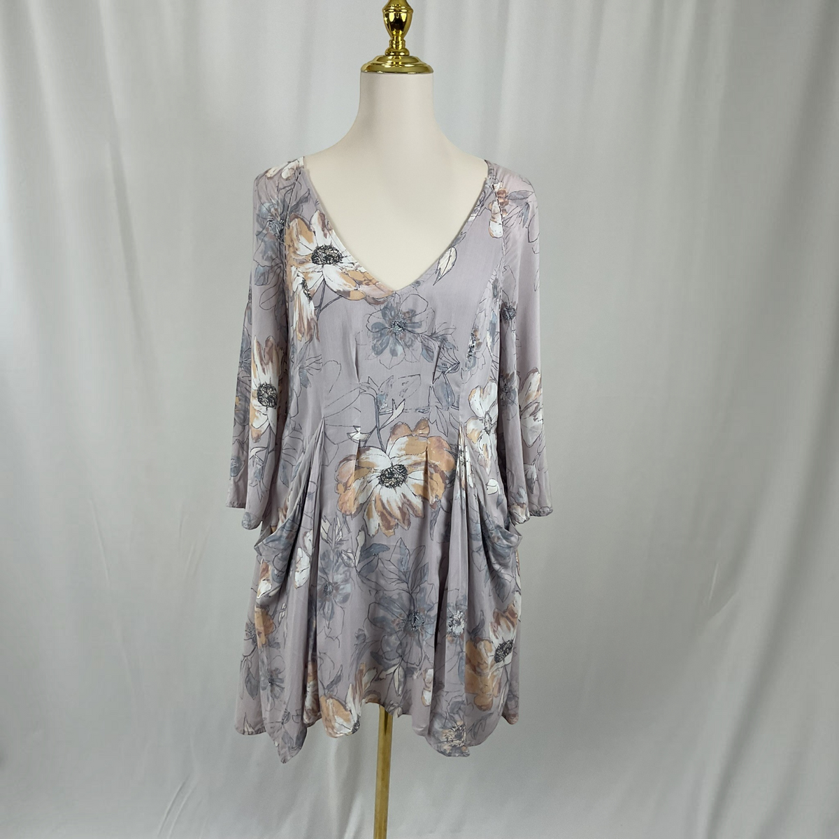 A three-quarter sleeve V-neck dress with a floral print in muted gray, beige, and white, displayed on a mannequin torso against a draped white fabric backdrop.
