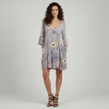 A woman modeling a light gray floral print dress with three-quarter sleeves and side pockets, paired with black strappy high-heeled sandals, against a neutral light gray background.