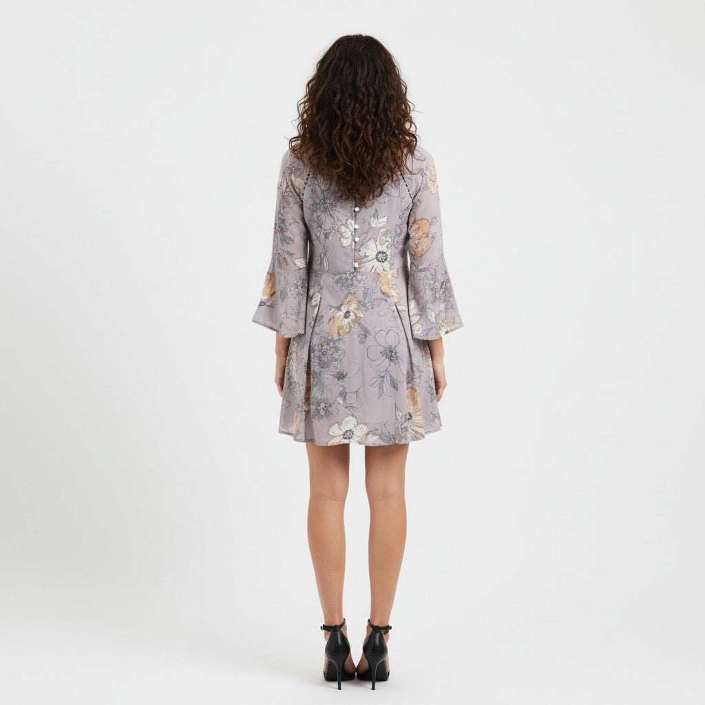 A woman viewed from behind wearing a short lavender-gray floral dress with three-quarter sleeves and black ankle-strap heels, standing against a plain light gray studio background.