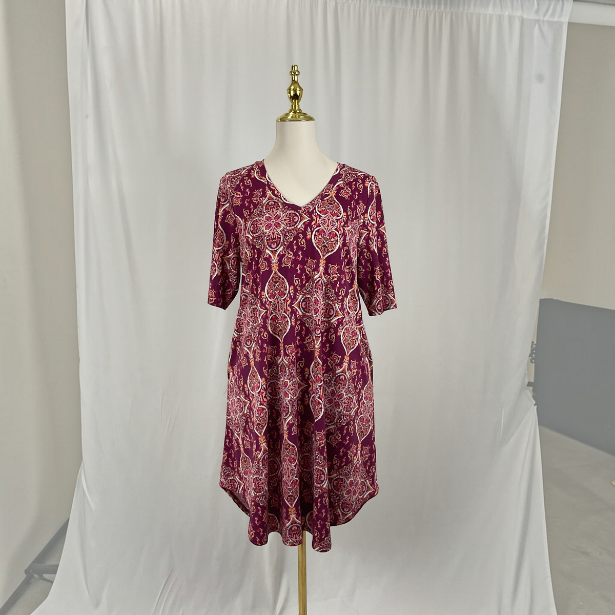 A short-sleeve V-neck dress with a purple and pink paisley pattern, displayed on a mannequin in front of a white fabric backdrop.