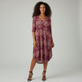 A woman modeling a short-sleeve V-neck tunic dress with a burgundy and beige paisley pattern, paired with black strappy high-heeled sandals, against a light gray studio background.