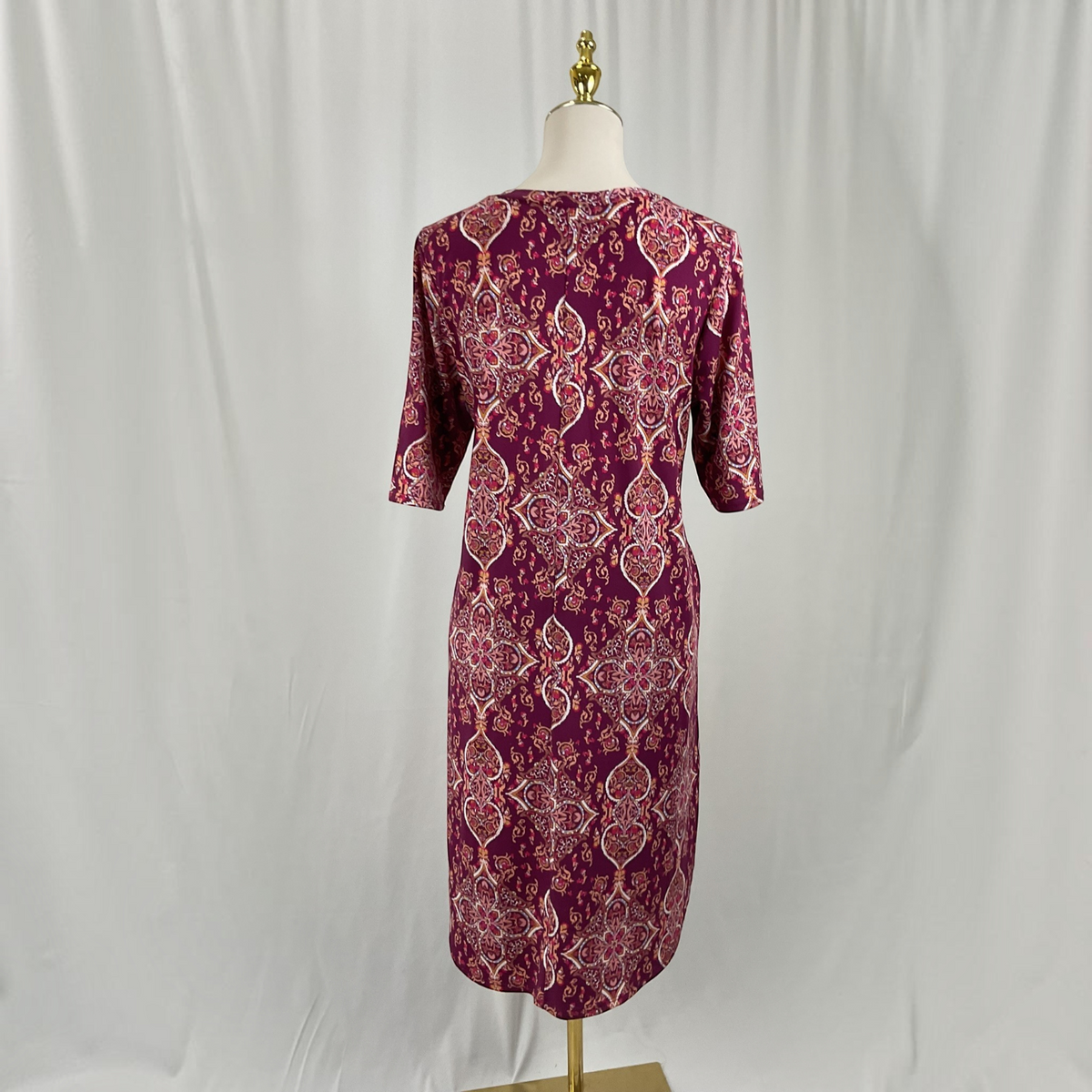 A knee-length dress displayed on a mannequin from the back, featuring a deep purple base with an intricate pink and white geometric and floral pattern.