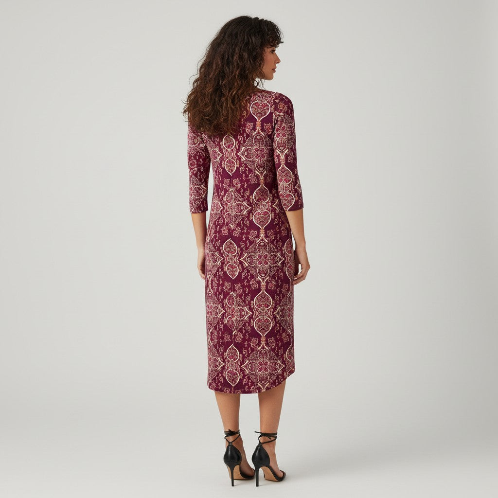 A woman stands with her back to the camera wearing a knee-length three-quarter sleeve dress with an intricate geometric and floral pattern in deep burgundy and beige. She is wearing black high-heeled sandals against a plain light gray studio background.