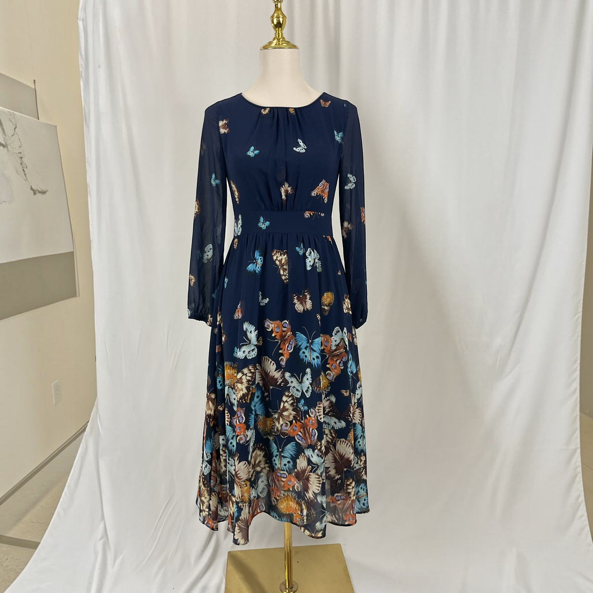 A navy blue long-sleeve midi dress with a colorful butterfly and floral print, displayed on a mannequin with a gold stand against a white fabric backdrop.
