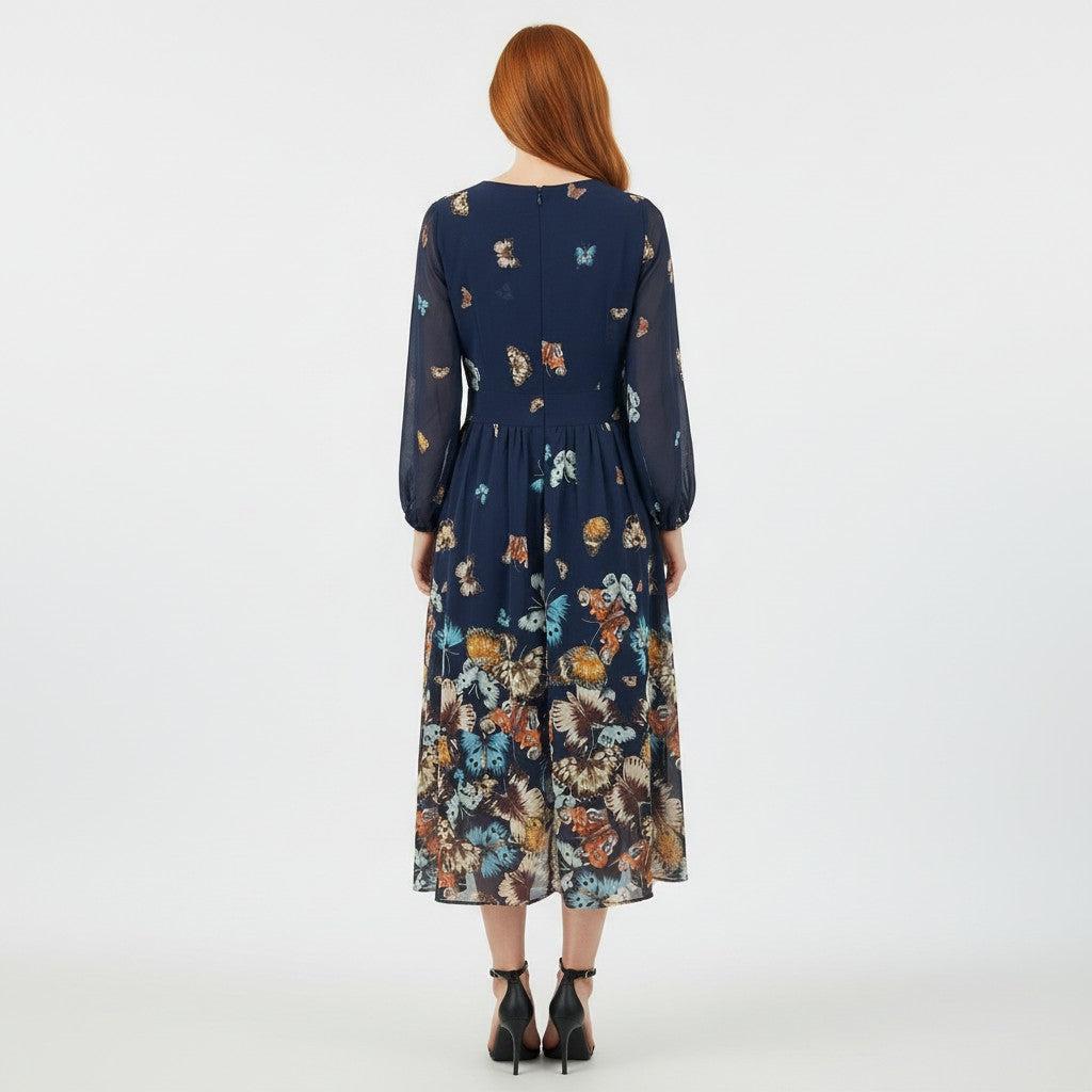 A woman viewed from the back wearing a navy midi dress with a colorful butterfly and floral print, paired with black ankle-strap heels, against a plain white studio background.
