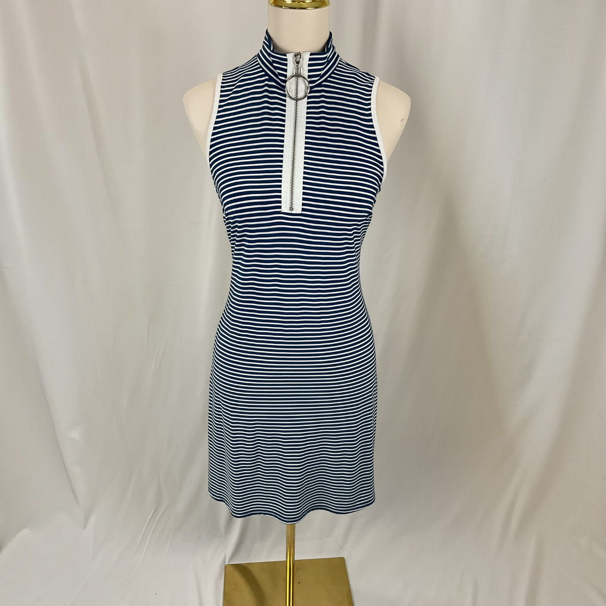 A sleeveless high-neck knit dress with thin navy and white horizontal stripes, featuring a white front zipper with a silver metal ring pull, displayed on a mannequin torso against a white draped fabric background.