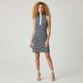 A woman modeling a sleeveless bodycon mini dress with thin navy and white horizontal stripes, a white mock neck collar, and white side piping, paired with white lace-up heeled sandals against a light gray studio background.