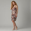 A woman modeling a sleeveless, one-shoulder ruffled dress with a pink, beige, and brown abstract print, paired with white high-heeled sandals, against a solid gray studio background.
