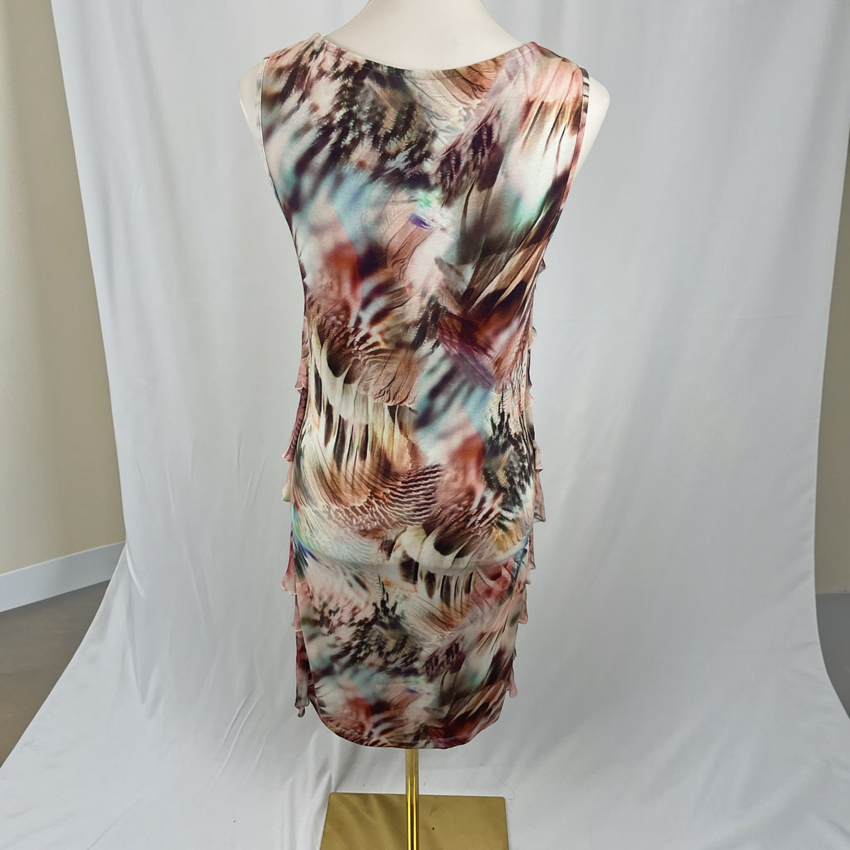 A sleeveless V-neck dress displayed on a mannequin torso, featuring an abstract feather-like print in brown, beige, pink, and muted blue tones, photographed against a white draped studio backdrop.