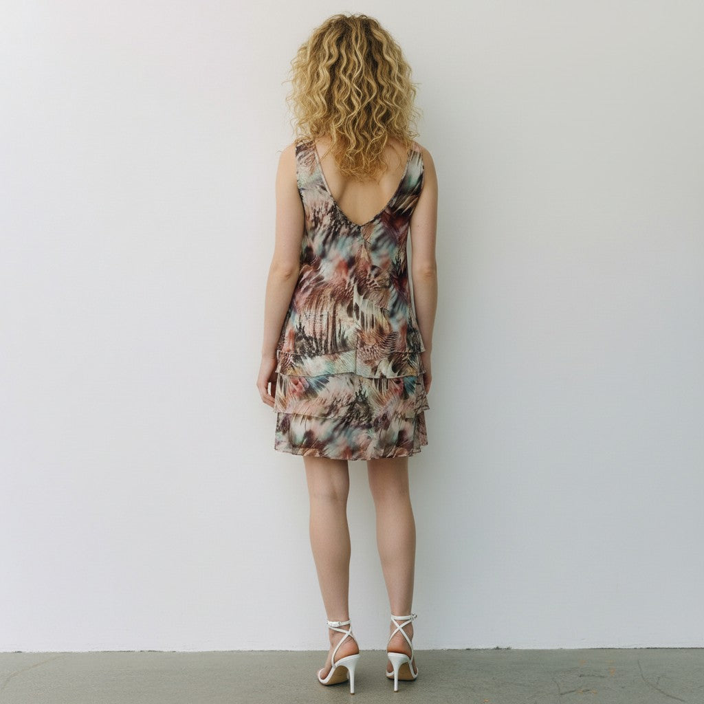 A woman with curly blonde hair is shown from behind wearing a sleeveless, tiered mini dress with a multicolor abstract print in shades of brown, beige, and teal. She is wearing white high-heeled sandals and standing against a plain white wall.