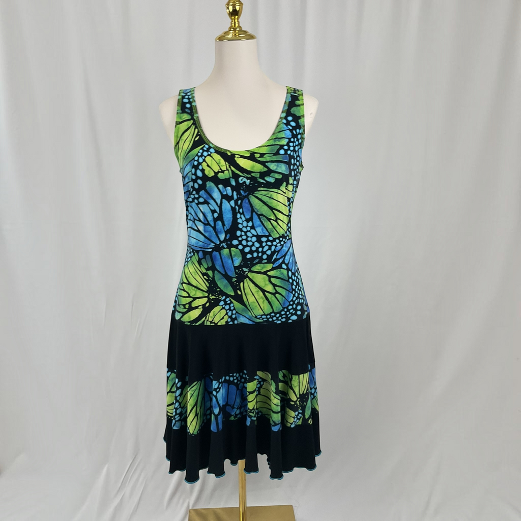 A sleeveless scoop-neck dress displayed on a mannequin against a white draped backdrop. The dress features a vibrant butterfly print in green, blue, and black, with a solid black tiered ruffle hem.