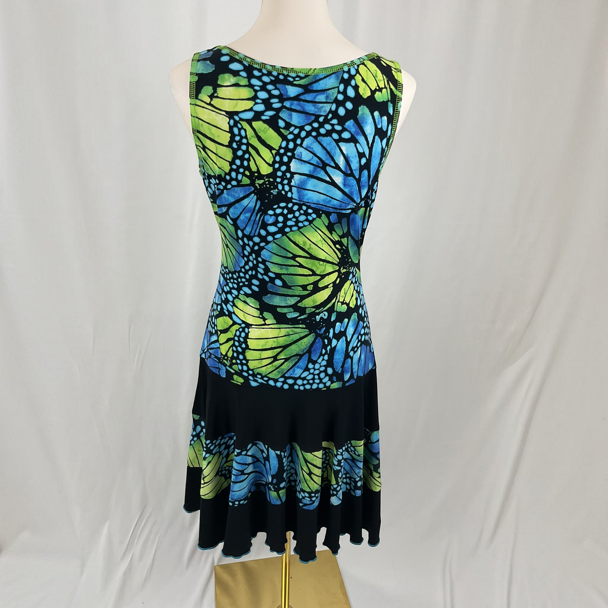 A sleeveless fit-and-flare dress displayed on a mannequin against a white draped backdrop. The dress features a vibrant butterfly wing print in blue, green, and black, with a solid black flared hem.