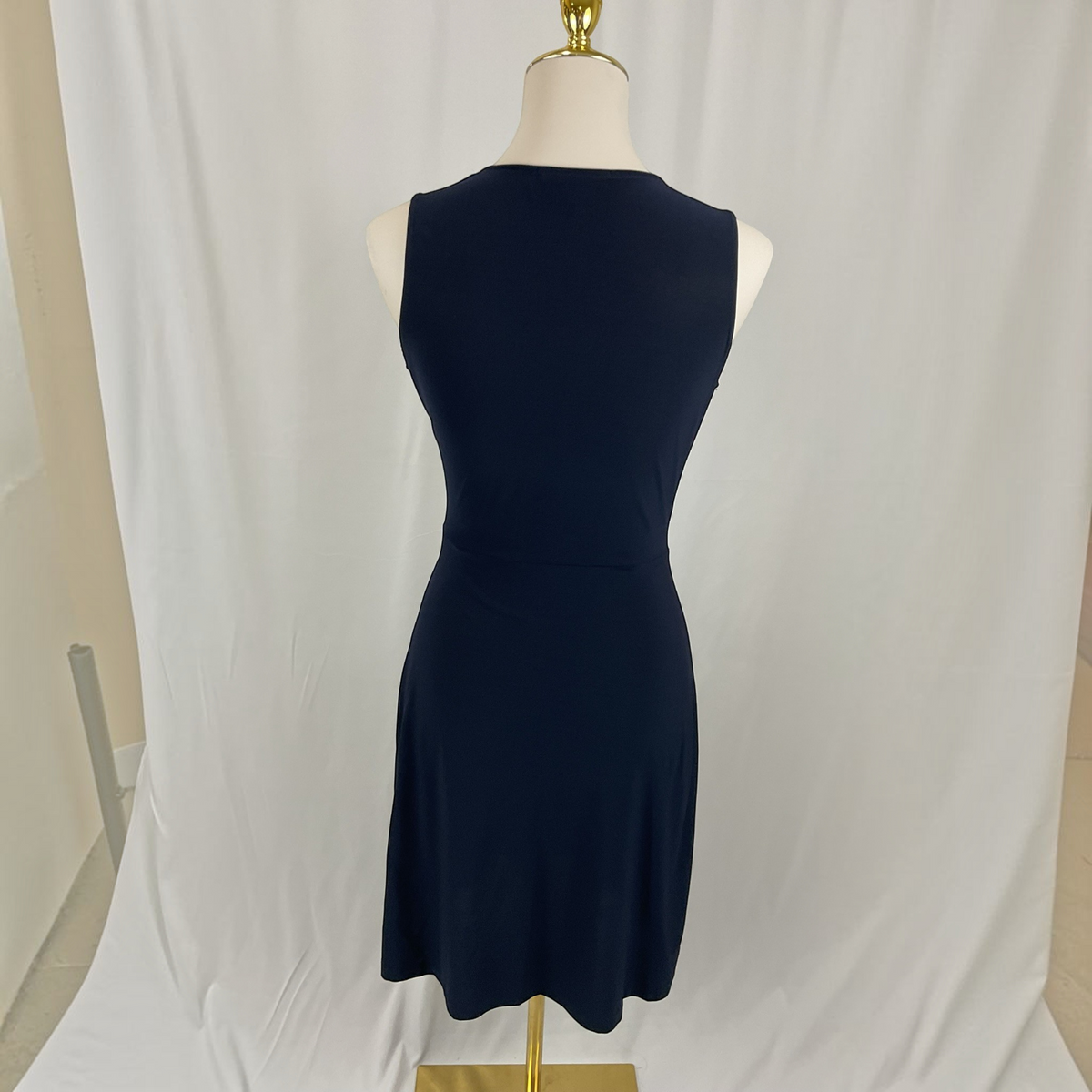 A sleeveless dark navy blue dress displayed on a mannequin torso with a gold stand, set against a draped white fabric backdrop.