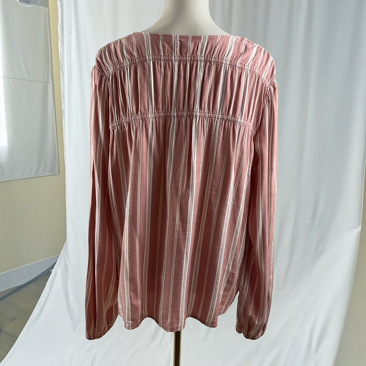 A long-sleeve blouse with vertical pink, white, and beige stripes, featuring a smocked bodice and elasticized cuffs, displayed on a mannequin torso against a white fabric backdrop.