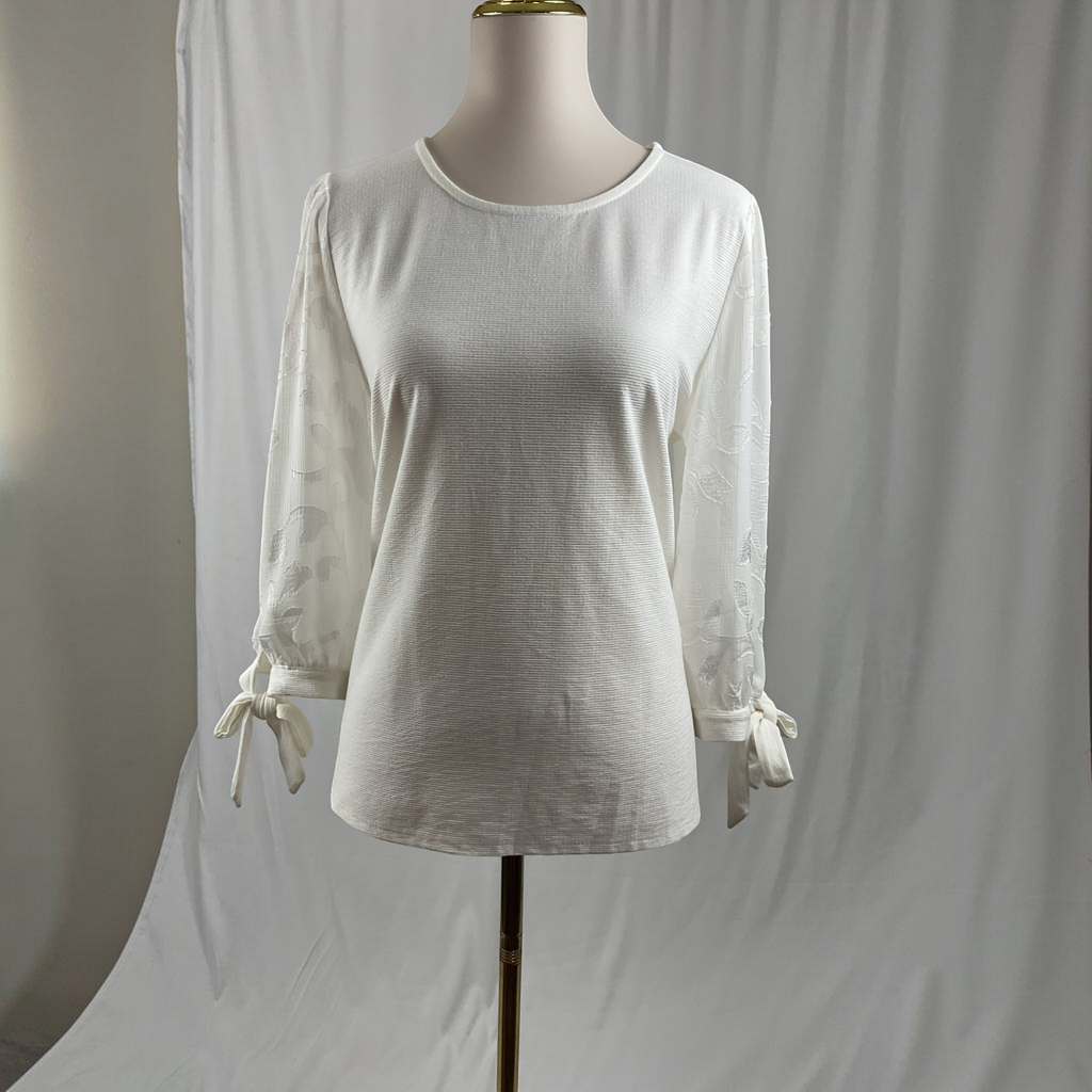 An off-white long-sleeve blouse with sheer patterned sleeves and tie details at the cuffs, displayed on a dress form against a white studio backdrop.