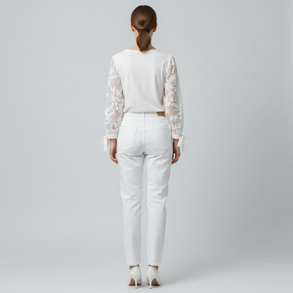 A woman viewed from behind wearing a white long-sleeve top with sheer floral lace sleeves and white straight-leg jeans, standing against a light gray studio background.