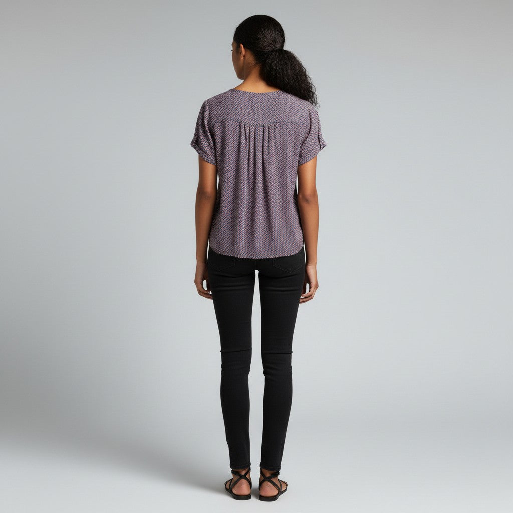 A woman viewed from the back wearing a short-sleeve top with a subtle purple and white pattern, paired with black skinny jeans and black strappy sandals, against a light gray studio background.