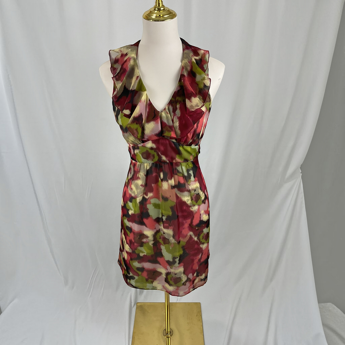 A sleeveless V-neck mini dress with a floral abstract print in burgundy, olive green, and beige, displayed on a white mannequin torso with a gold stand against a white fabric backdrop.