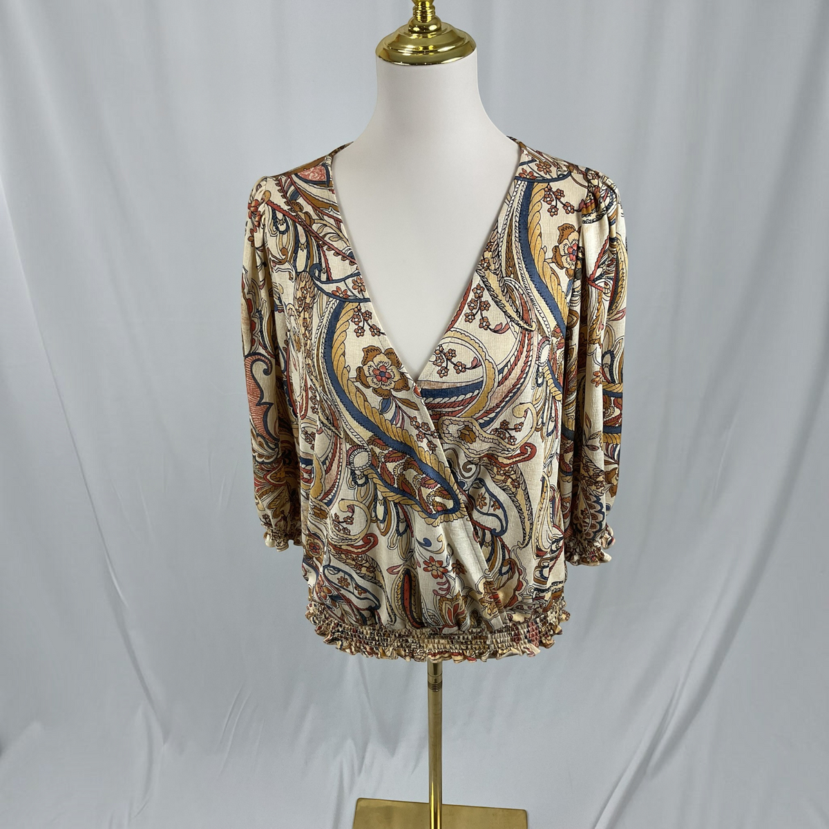 A woman modeling a beige and brown paisley patterned blouse with dark navy skinny jeans and black strappy sandals, standing against a light gray studio background.