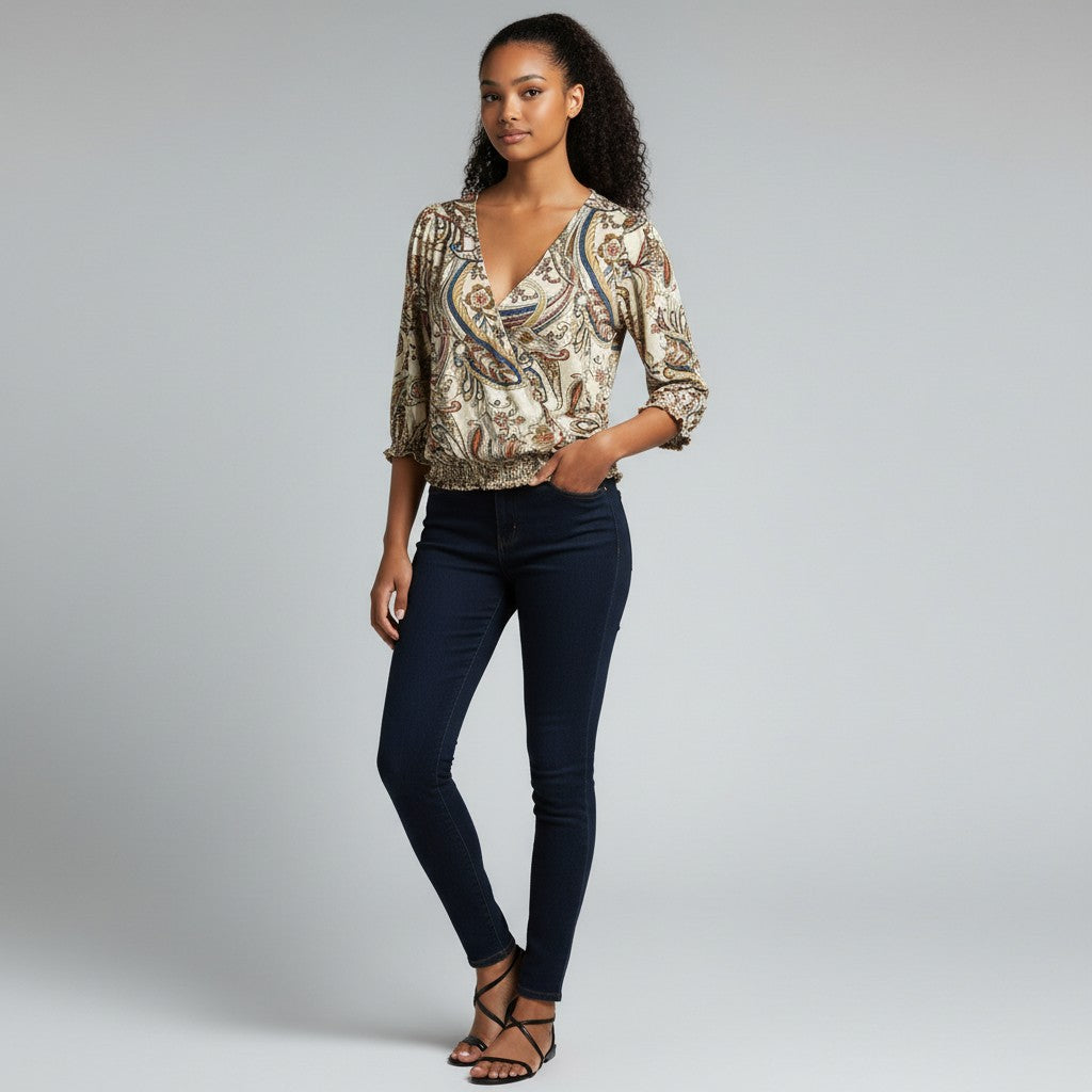 A woman modeling a beige and brown paisley patterned blouse with dark navy skinny jeans and black strappy sandals, standing against a light gray studio background.