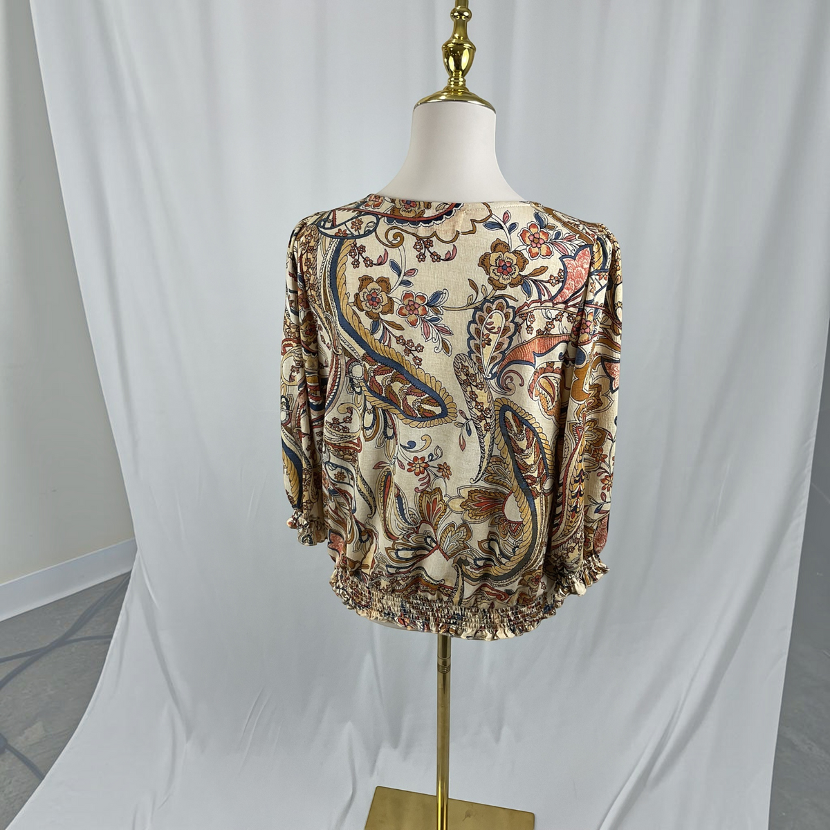 A woman modeling a beige and brown paisley patterned blouse with dark navy skinny jeans and black strappy sandals, standing against a light gray studio background.