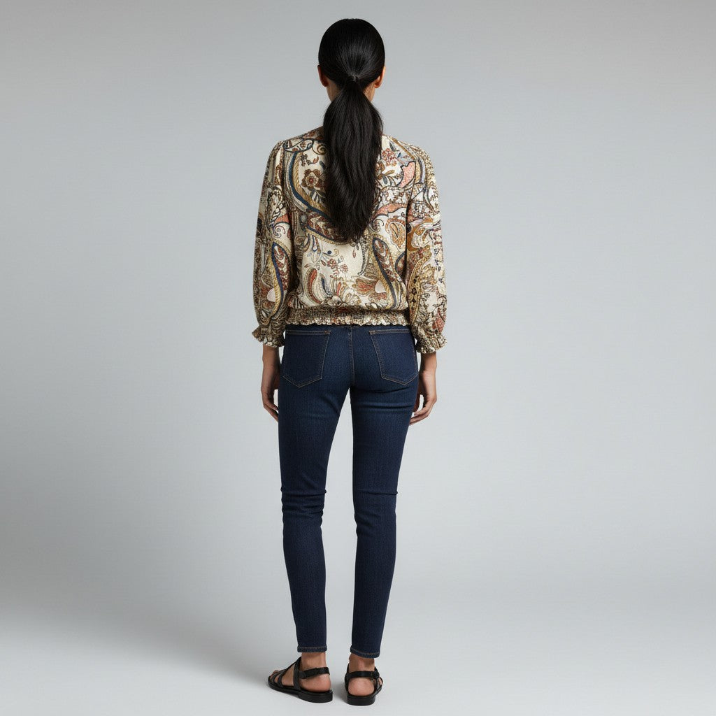 A woman modeling a beige and brown paisley patterned blouse with dark navy skinny jeans and black strappy sandals, standing against a light gray studio background.