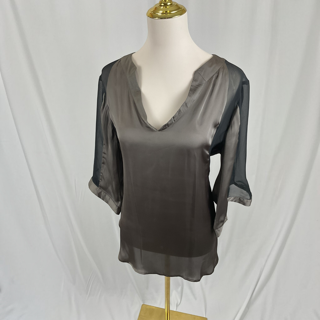 A woman modeling a loose, semi-sheer taupe-gray blouse with sheer three-quarter sleeves, paired with dark blue skinny jeans and black sandals, against a light gray studio background.