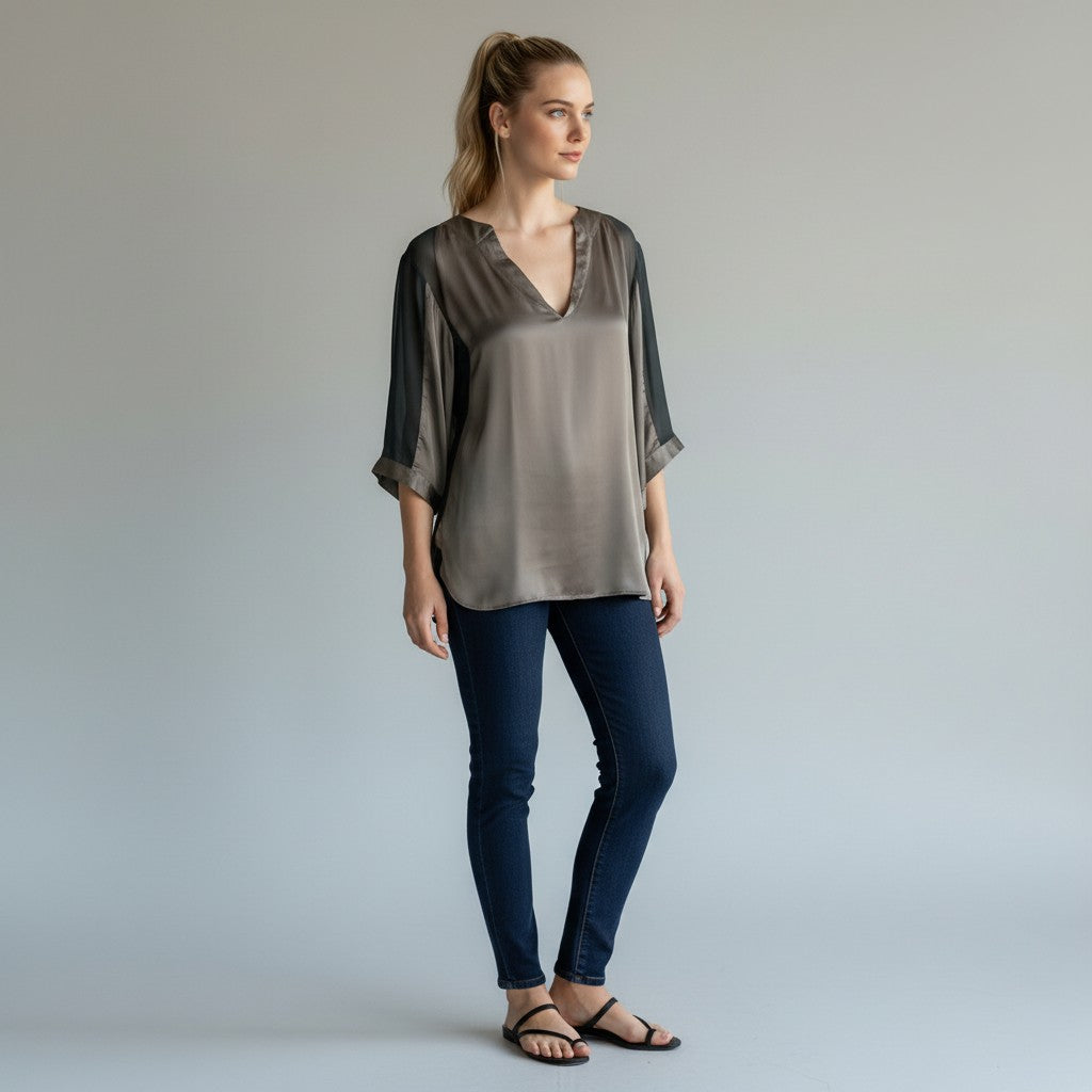 A woman modeling a loose, semi-sheer taupe-gray blouse with sheer three-quarter sleeves, paired with dark blue skinny jeans and black sandals, against a light gray studio background.