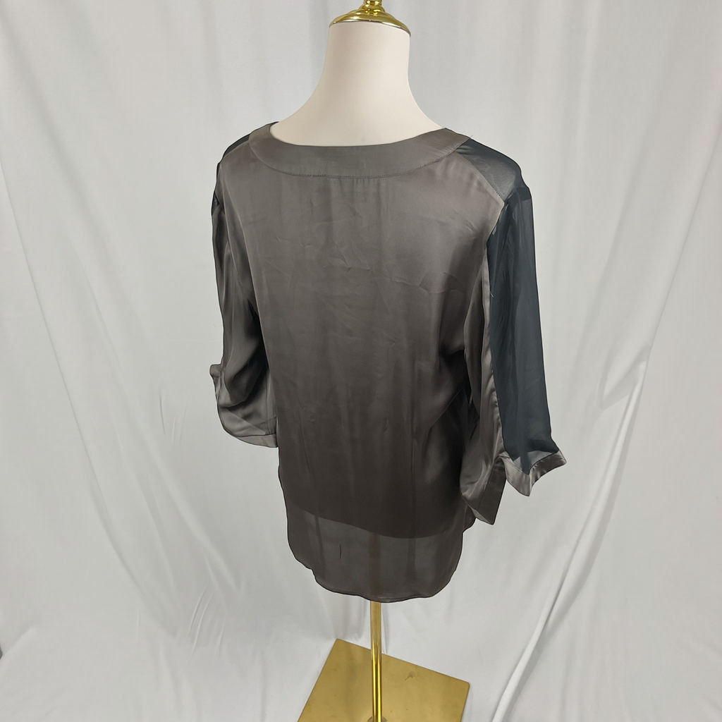 A woman modeling a loose, semi-sheer taupe-gray blouse with sheer three-quarter sleeves, paired with dark blue skinny jeans and black sandals, against a light gray studio background.