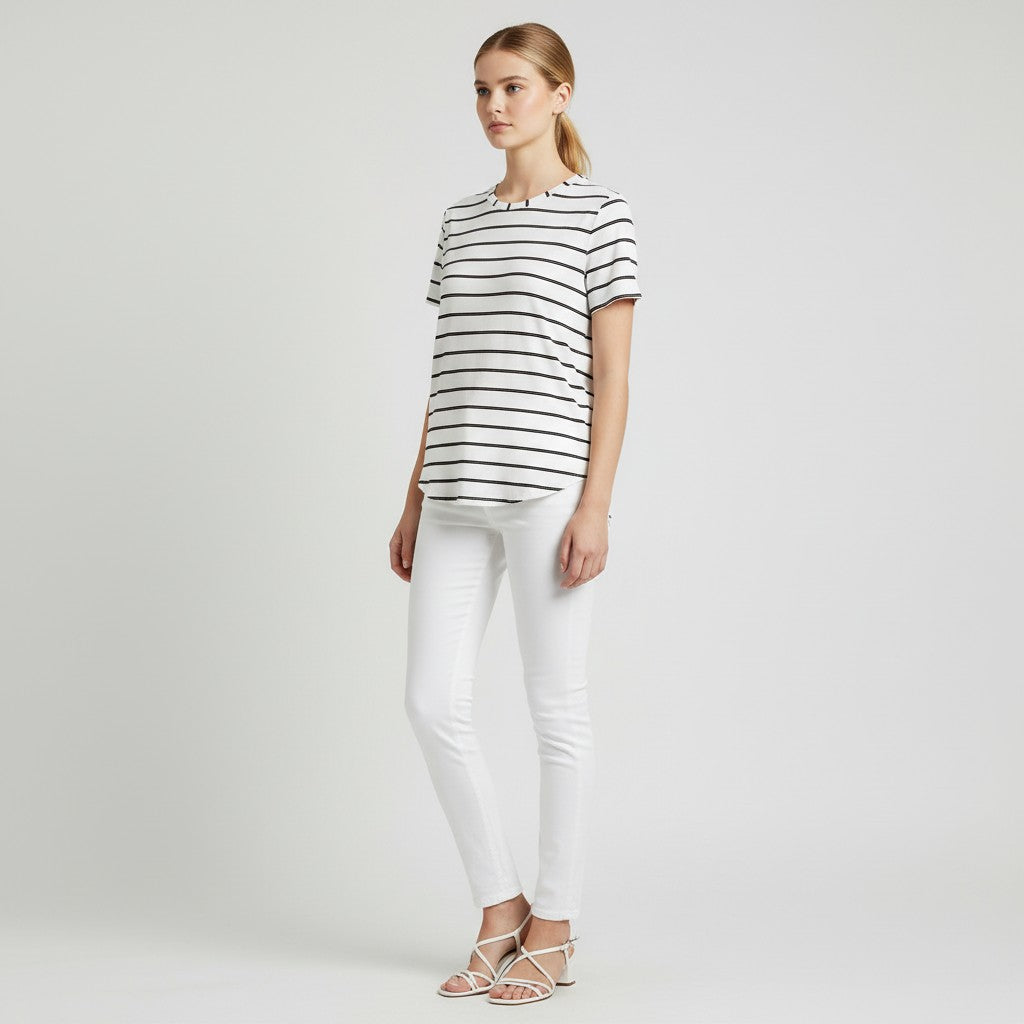 A woman modeling a short-sleeve crewneck t-shirt with thin black horizontal stripes on a white base, paired with white skinny jeans and white strappy flat sandals, against a light gray studio background.