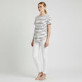 A woman modeling a short-sleeve crewneck t-shirt with thin black horizontal stripes on a white base, paired with white skinny jeans and white strappy flat sandals, against a light gray studio background.