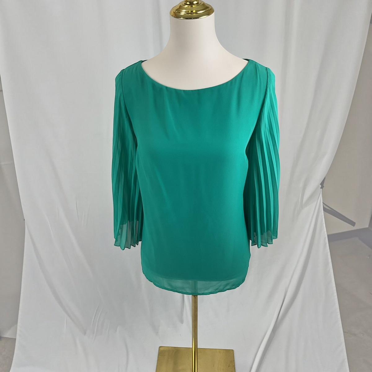 A woman modeling a vibrant green pleated three-quarter sleeve top paired with white skinny jeans and white heeled sandals, standing against a neutral gray studio background.