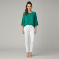 A woman modeling a vibrant green pleated three-quarter sleeve top paired with white skinny jeans and white heeled sandals, standing against a neutral gray studio background.