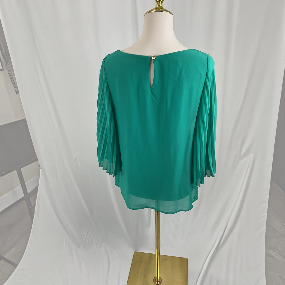 A woman modeling a vibrant green pleated three-quarter sleeve top paired with white skinny jeans and white heeled sandals, standing against a neutral gray studio background.
