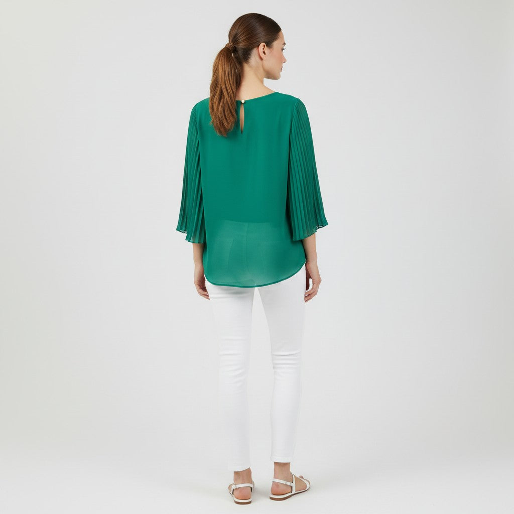 A woman viewed from the back wearing a green pleated long-sleeve blouse and white pants, standing against a plain light gray studio background.