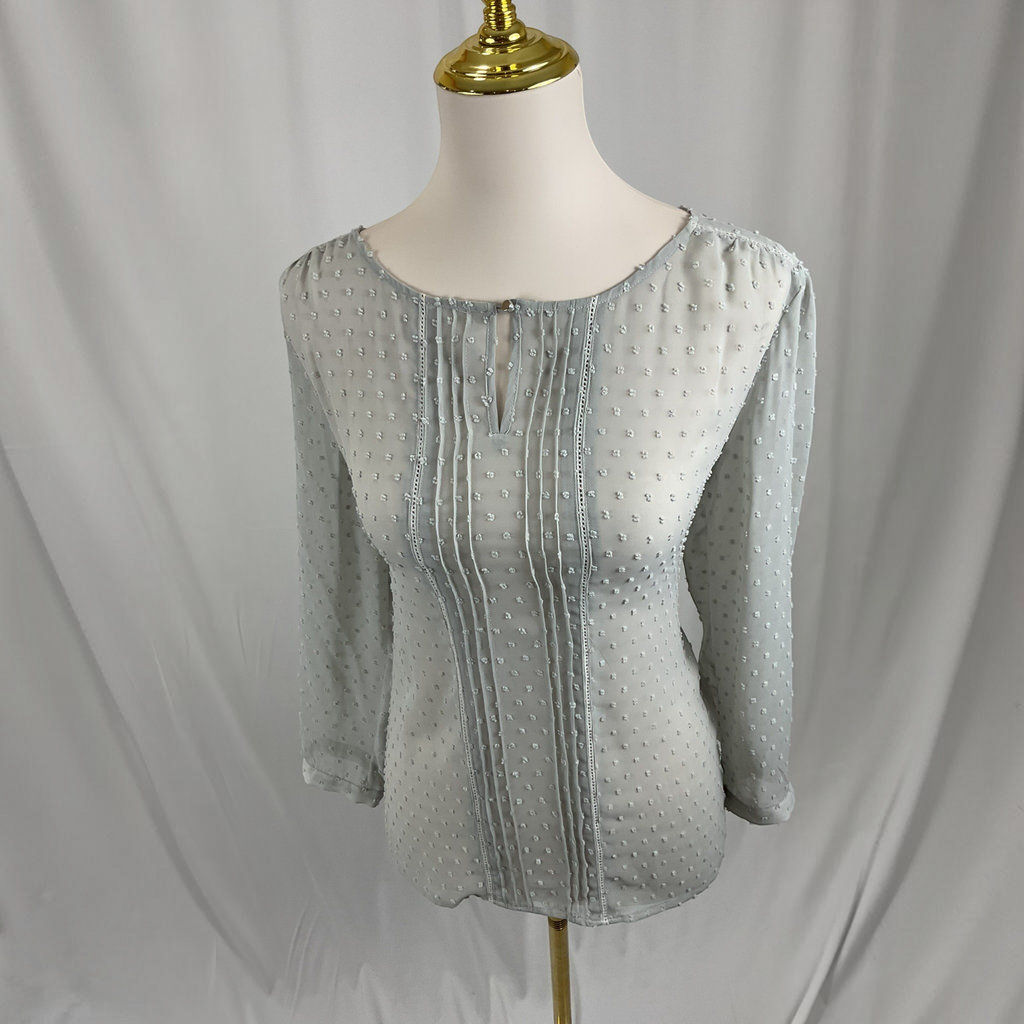 A light gray three-quarter sleeve blouse with a textured dotted pattern and vertical pleated detailing on the front, displayed on a cream mannequin torso with a gold neck cap against a draped white fabric backdrop.