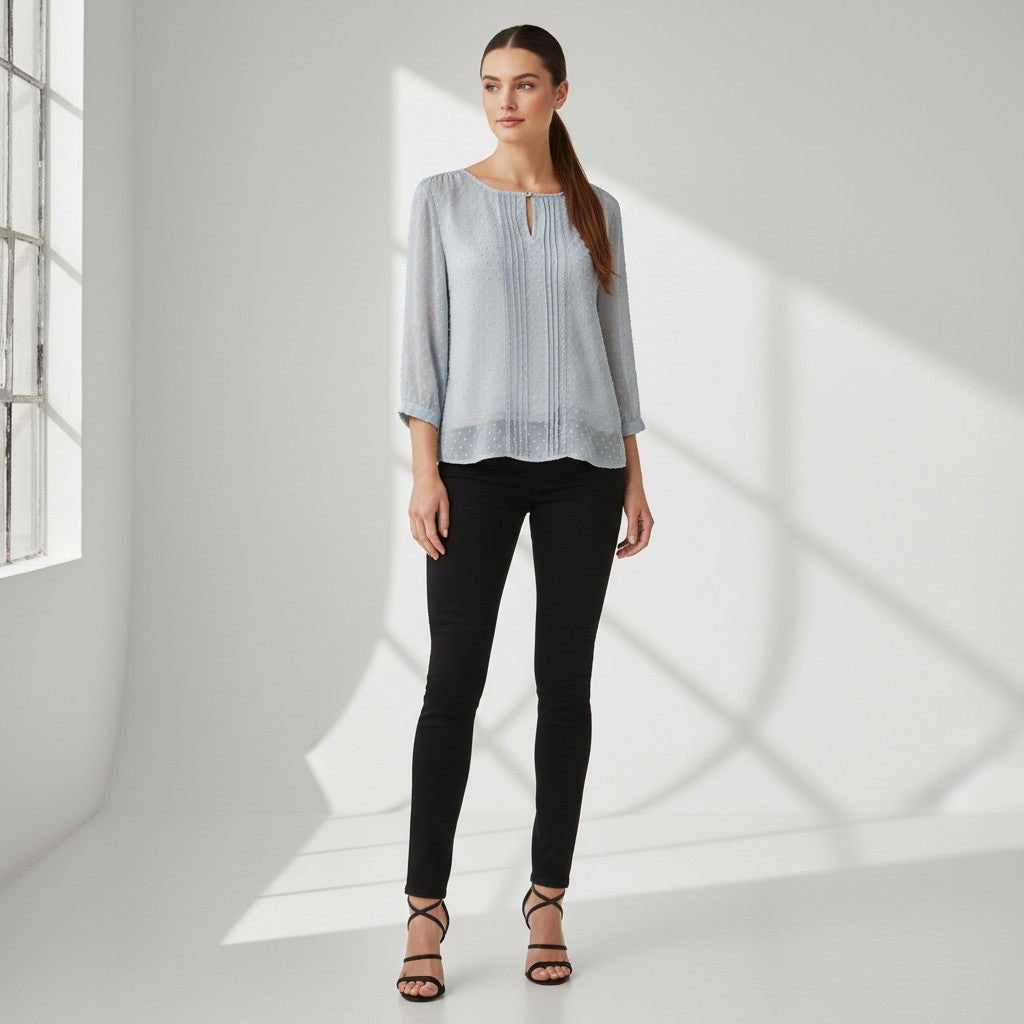 A woman stands in a bright white studio wearing a light blue long-sleeve top with a split neckline, black skinny pants, and black strappy heels.