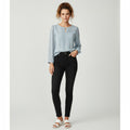 A woman modeling a light blue long-sleeve blouse with a keyhole cutout and black skinny jeans, standing against a light gray studio background.