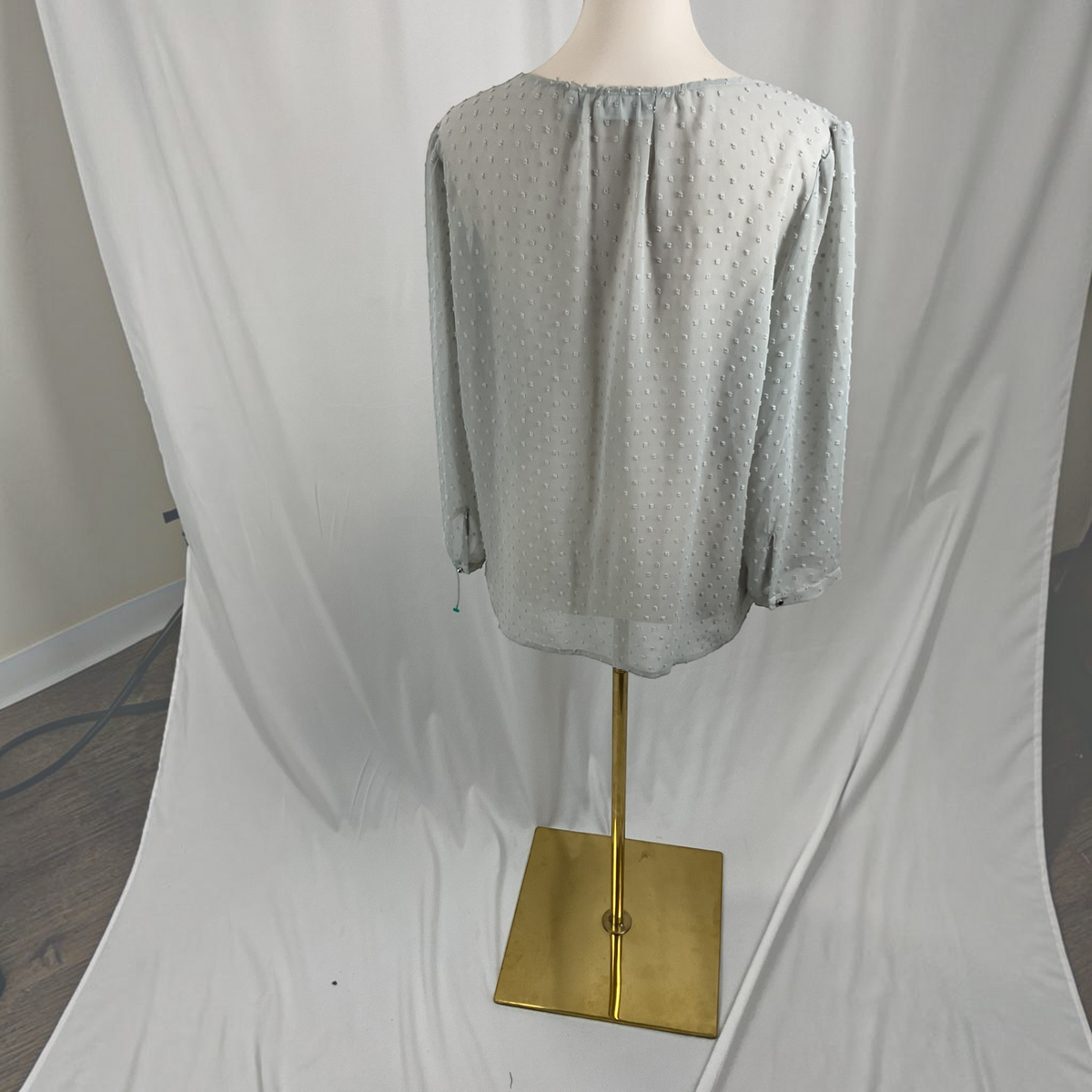 A light sage green long-sleeve blouse with a subtle dotted texture, displayed on a mannequin torso with a gold stand against a draped white fabric backdrop.