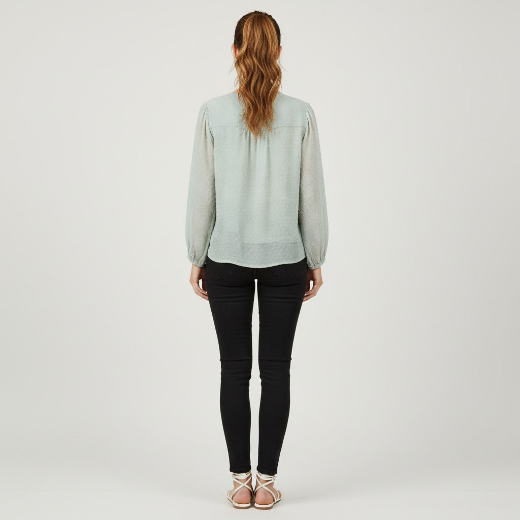 A woman viewed from behind wearing a light sage green long-sleeve blouse, black skinny jeans, and beige strappy sandals against a plain light gray studio background.