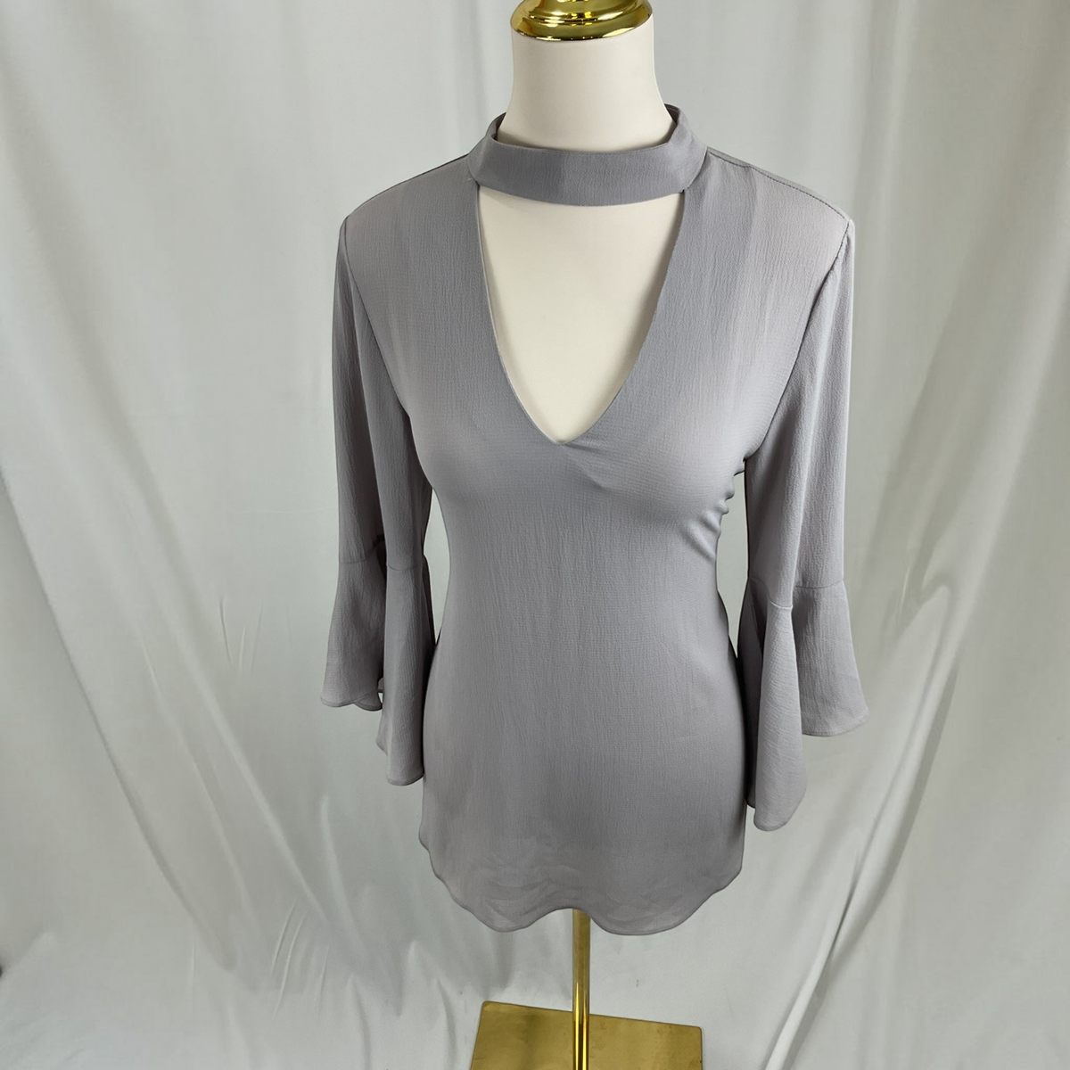 A light gray three-quarter sleeve blouse with a keyhole cutout and choker-style collar, displayed on a cream mannequin with a gold stand against a white draped fabric backdrop.