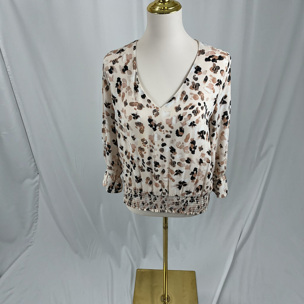 A women's long-sleeve blouse with a smocked waist and a brown, black, and beige floral pattern on a cream background, displayed on a white mannequin torso with a gold stand against a light gray fabric backdrop.