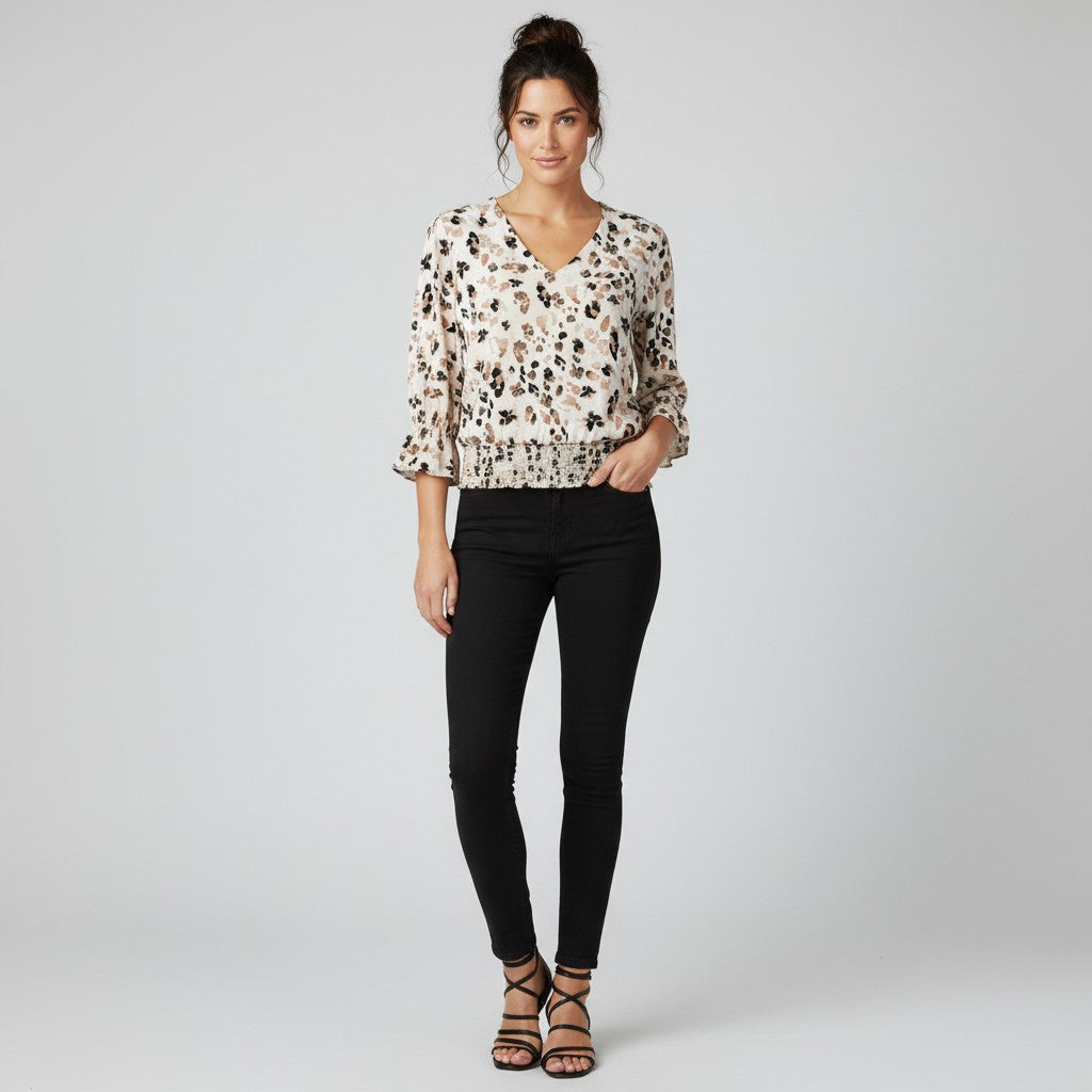 A woman modeling a cream-colored blouse with a brown and black abstract pattern, paired with black skinny jeans and brown strappy heels, against a light gray studio background.