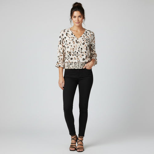 A woman modeling a cream-colored blouse with a brown and black abstract pattern, paired with black skinny jeans and brown strappy heels, against a light gray studio background.