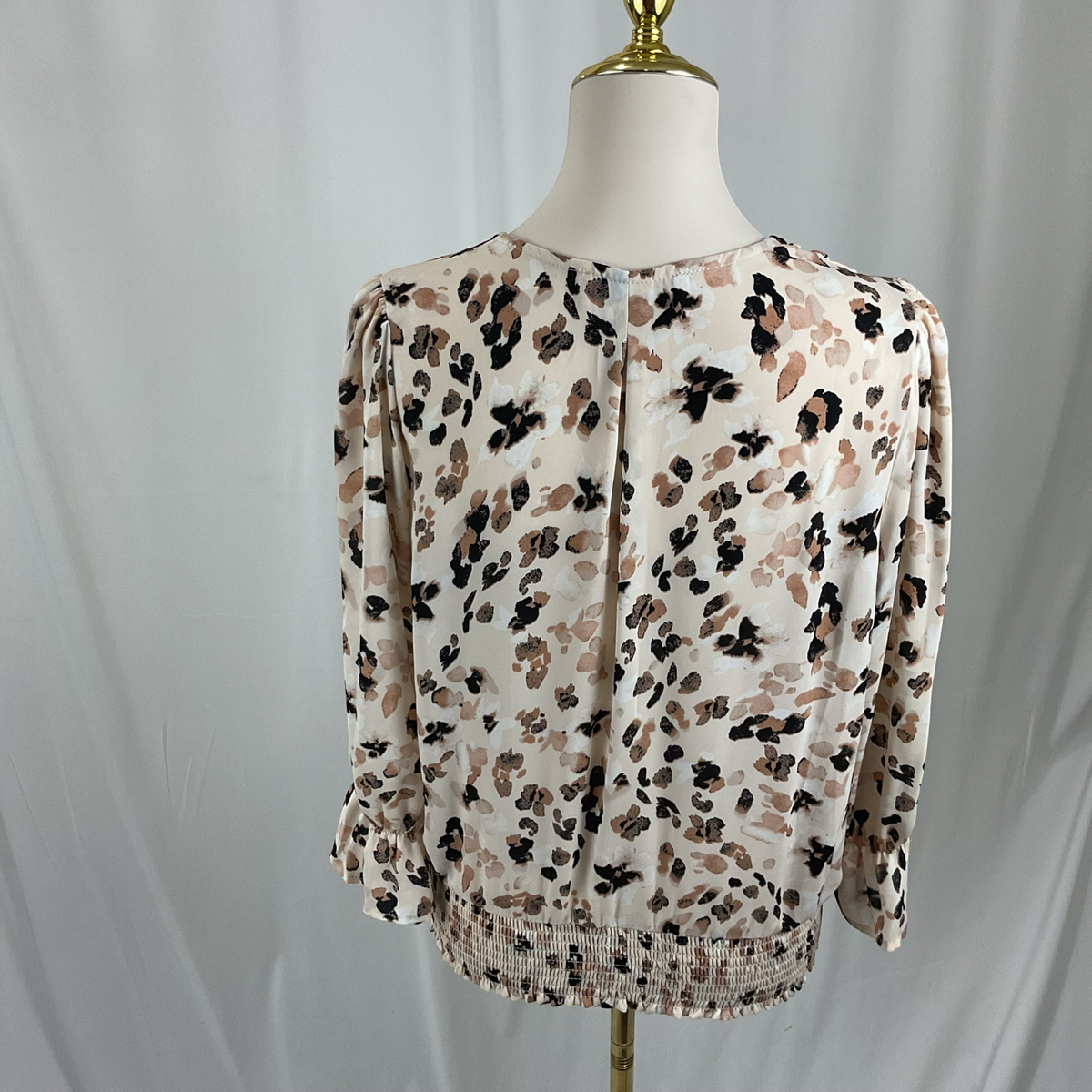 A three-quarter sleeve blouse with a beige, brown, and black abstract floral pattern, displayed on a mannequin torso against a white draped fabric background.