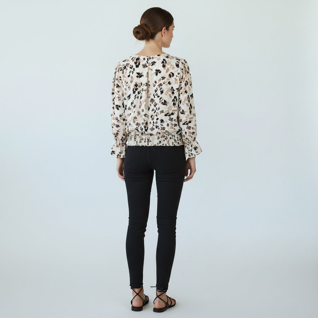 A woman viewed from behind wearing a beige long-sleeve blouse with a black and brown abstract animal print, paired with black skinny pants and black strappy sandals, against a light gray studio background.