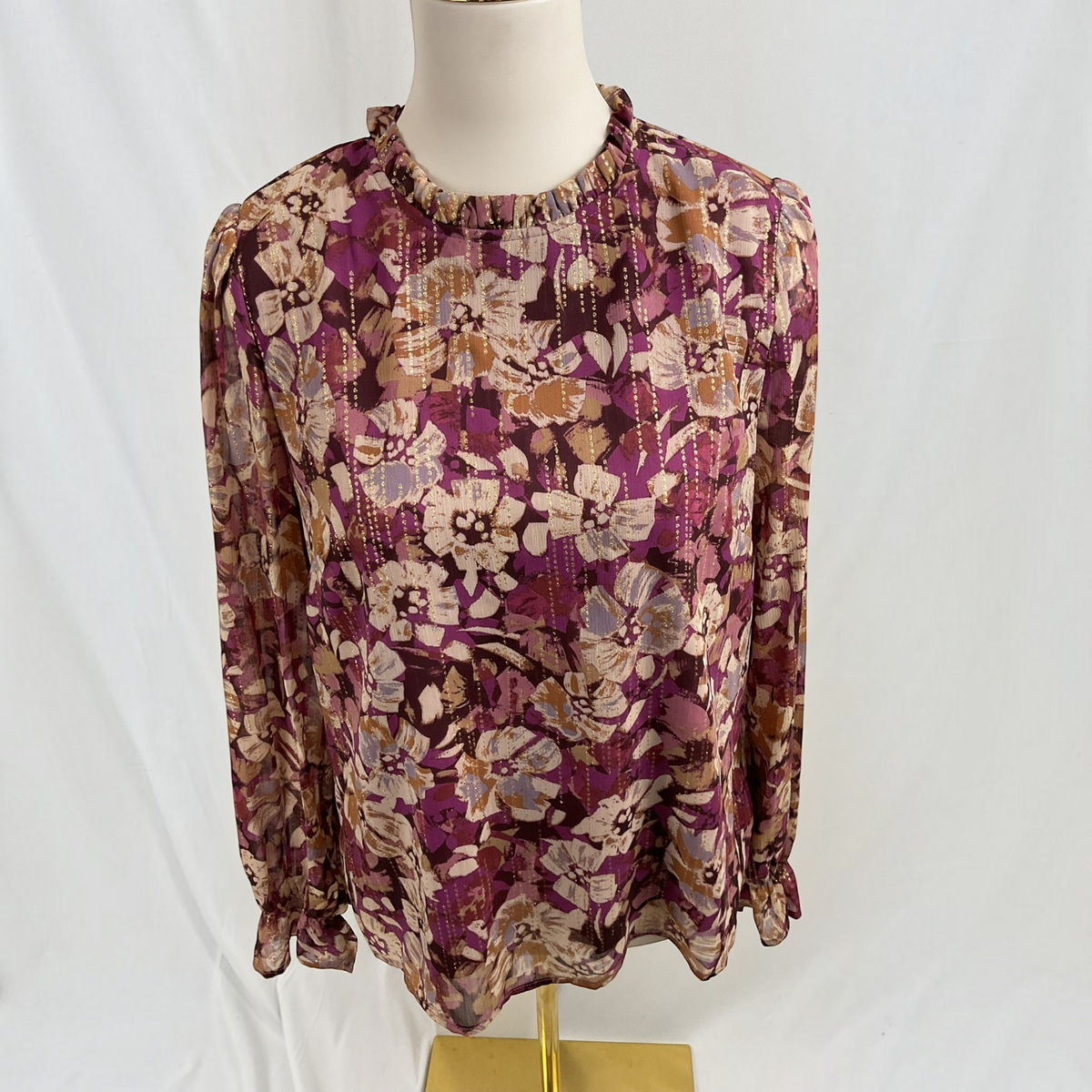 A long-sleeve blouse with a ruffled high neck and an all-over floral pattern, displayed on a mannequin torso against a white fabric backdrop.
