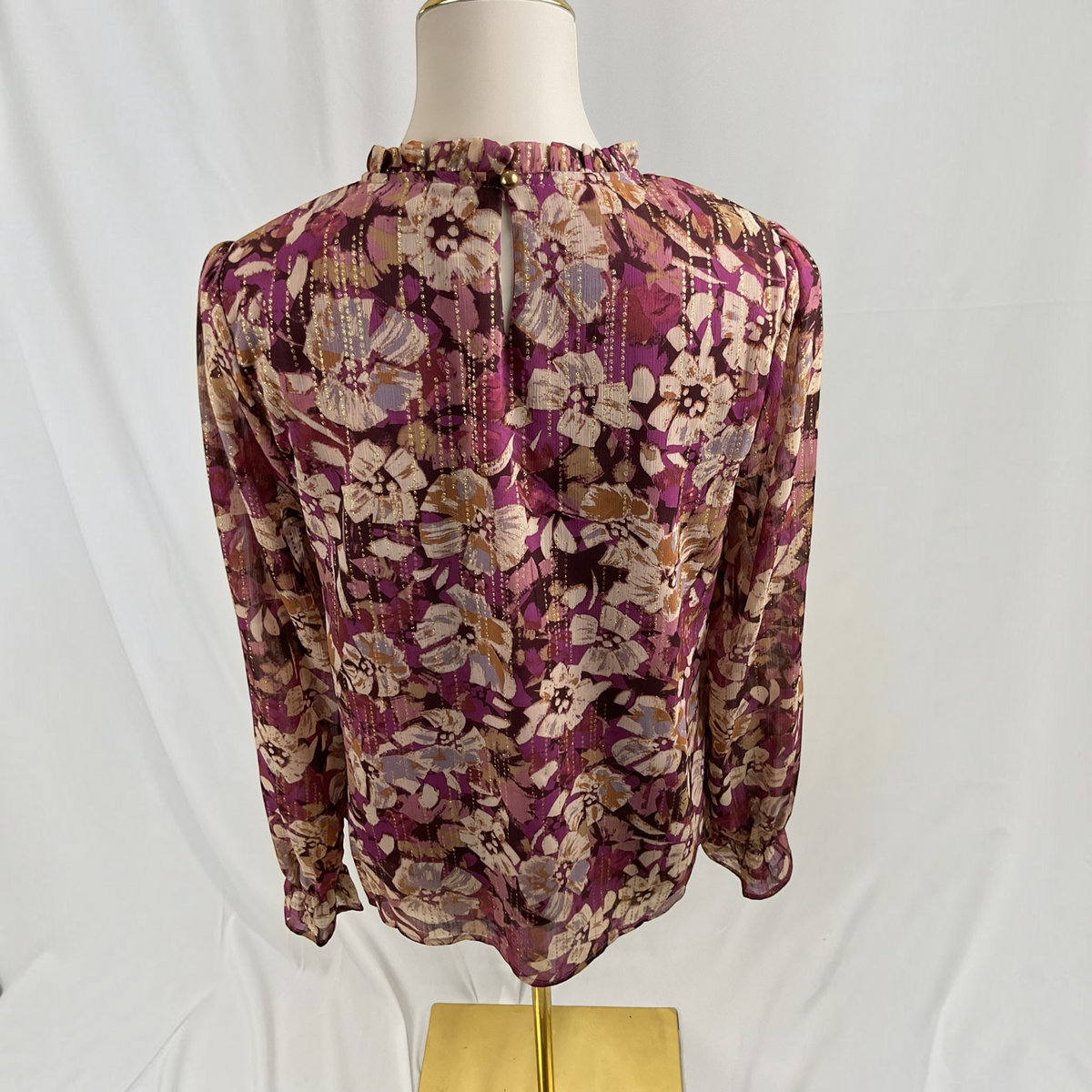A long-sleeve floral blouse displayed on a mannequin torso against a white draped backdrop. The blouse features a dense floral pattern in shades of purple, beige, brown, and gray, with a ruffled neckline.