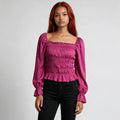 A woman with red hair wearing a magenta smocked long-sleeve top with a square neckline and black jeans, standing against a light gray studio background.