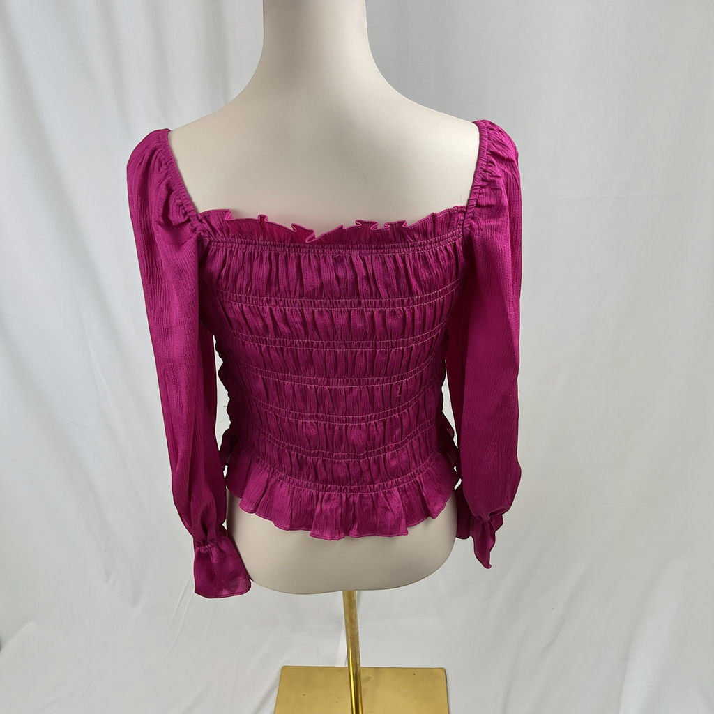 A magenta off-the-shoulder smocked blouse with long sleeves, displayed on a cream mannequin torso with a gold stand against a white draped backdrop.